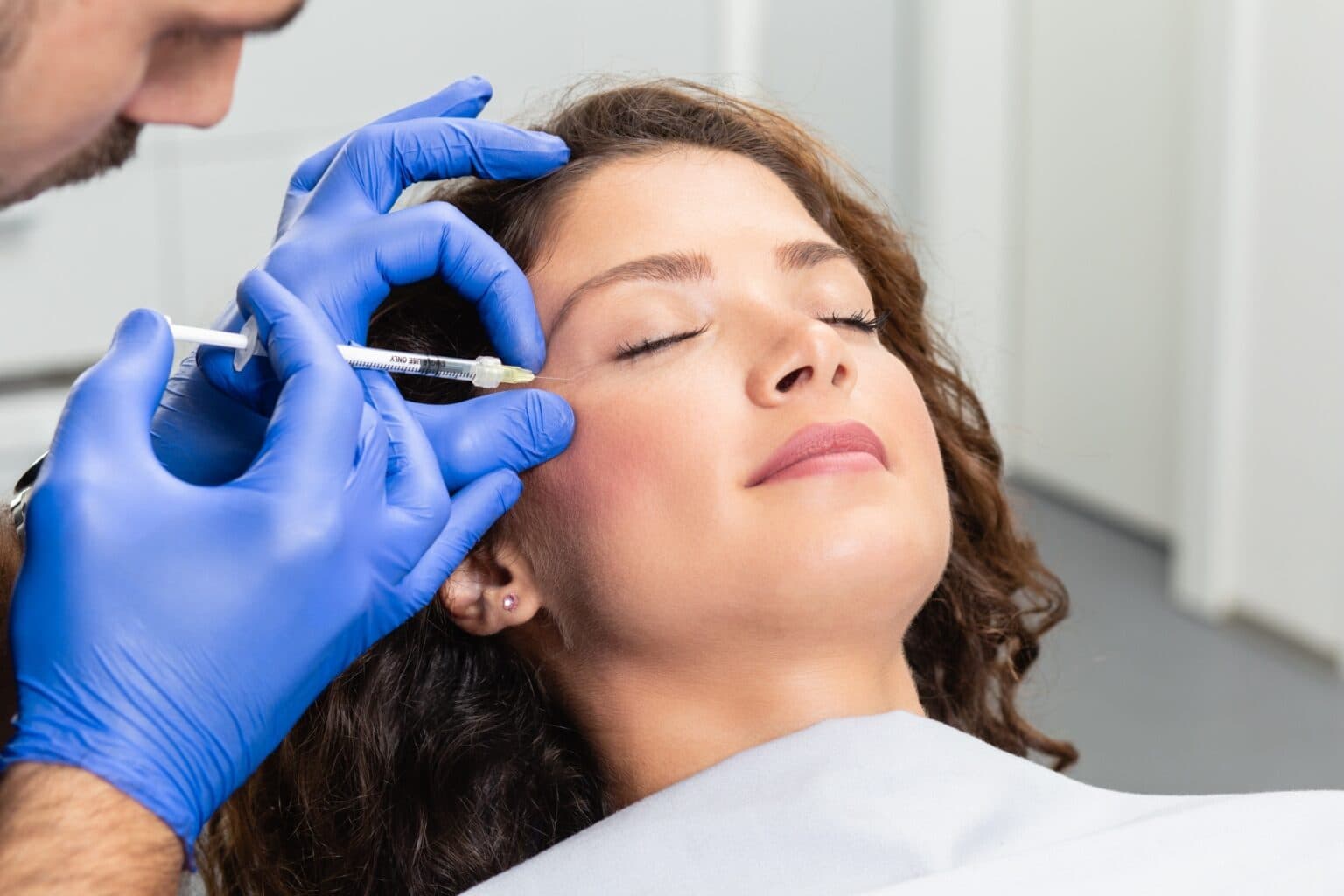 A clinician giving a small injection at the outer brow, the patient reclined with her eyes closed